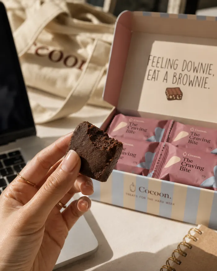 A hand holding a Craving Bite, the open box behind on a desk